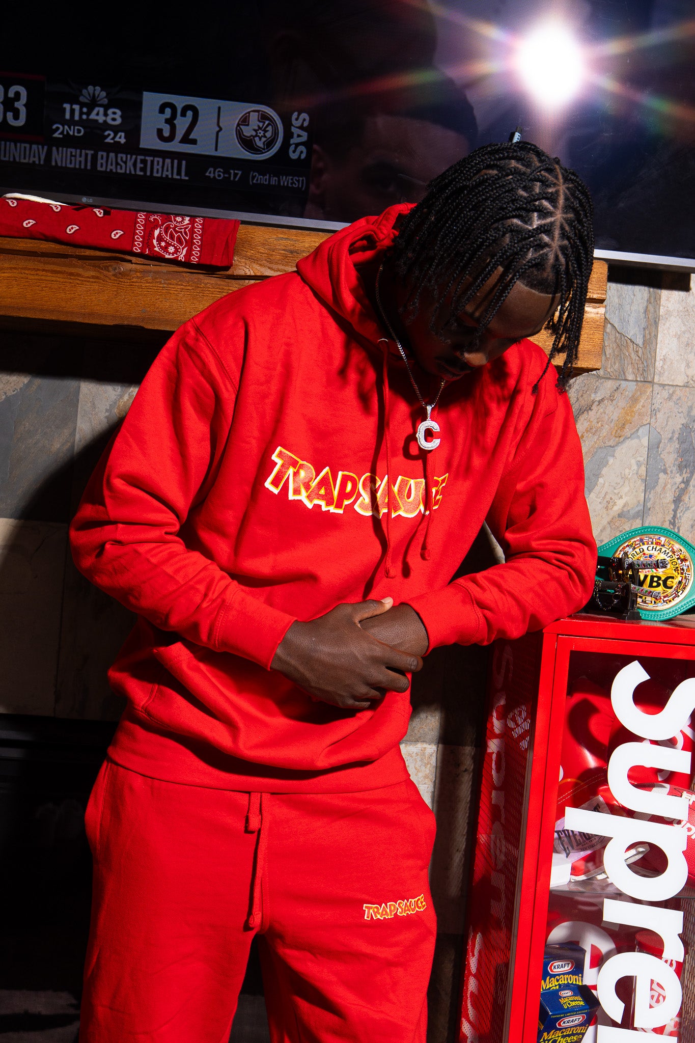 Person wearing a red trap sauce hoodie and pants standing in front of a TV displaying a basketball game, with a 'Supreme' box and various items in the background.