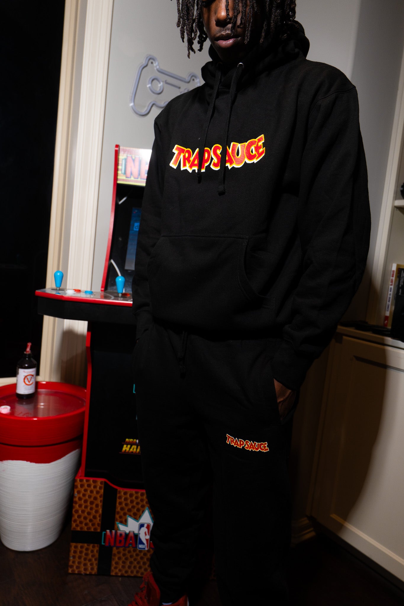 Person wearing a TSF chain and black sweatshirt with trap sauce logo in a room with nba jams retro game equipment.