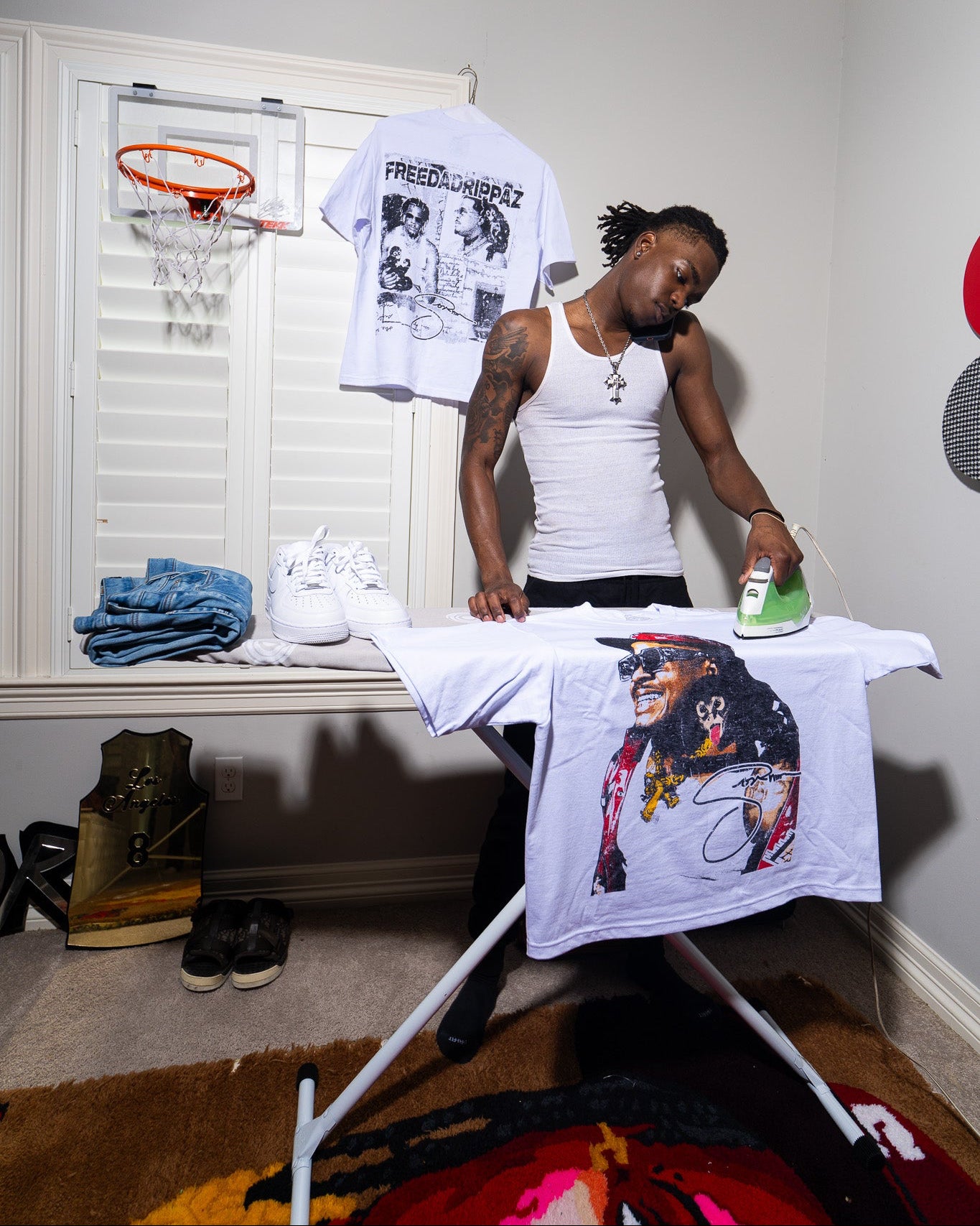 Person ironing a t-shirt with a Sosamann design in a room with a basketball hoop and sports-themed decor.