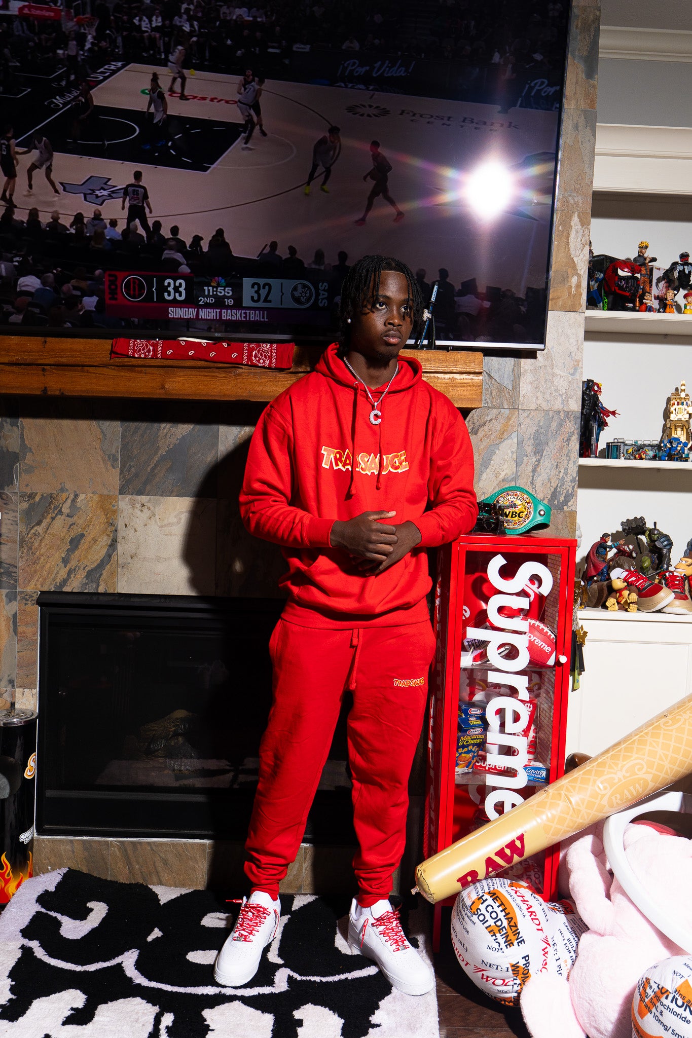 Person wearing a red trap sauce hoodie and pants standing in front of a TV displaying a basketball game, with a 'Supreme' box and various items in the background.